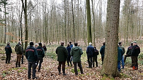 Eine Gruppe von Menschen steht in einem Waldstück. Es ist Winter, die Bäume tragen kein Laub.