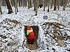 Eine Person mit rot-gelber Arbeitskleidung steht in einem Loch, das in den Waldboden gegraben wurde. Der Waldboden ist schneebedeckt. 