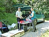 Auf einem Waldweg steht ein Bus, vor dem ein Tisch aufgebaut ist. Darauf bereiten ein Mann und eine Frau verschiedene Proben von Eschen für den Transport vor. Diese können anschließend in die neben dem Tisch stehenden Styroporboxen gepackt werden. 